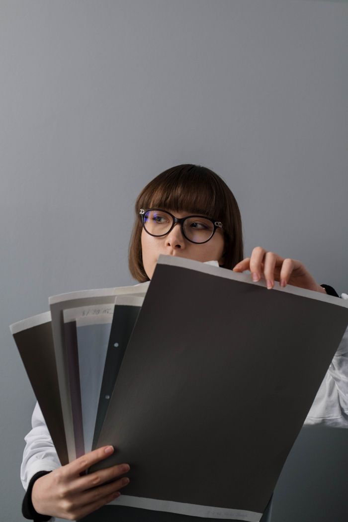 who-we-are A woman wearing glasses examines various paper samples, offering a minimalistic and modern aesthetic.